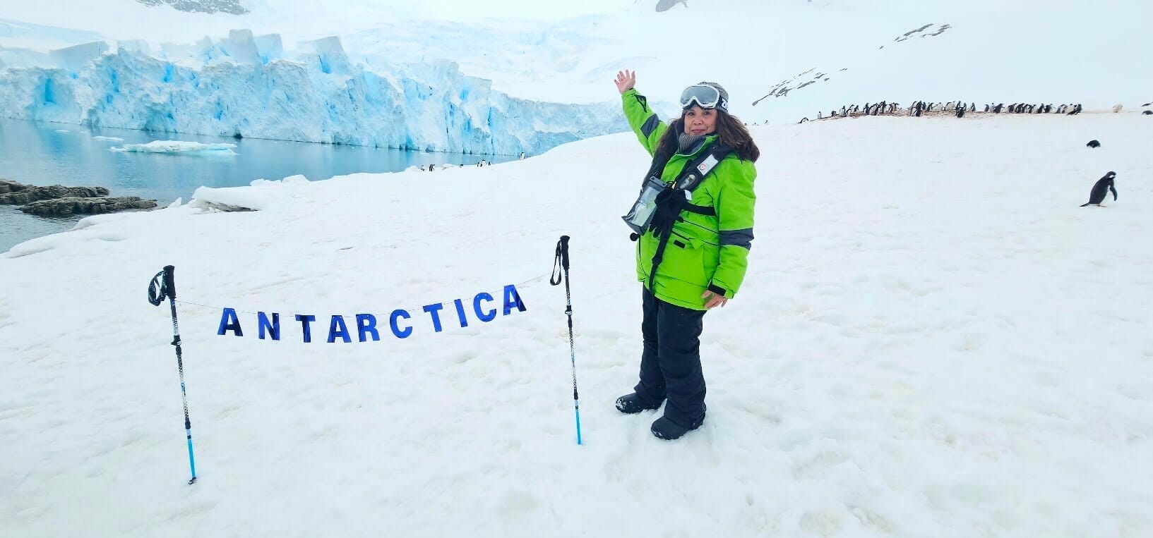 Explorer in green jacket on Antarctica snowfield with penguins, icebergs, and "ANTARCTICA" sign in foreground.