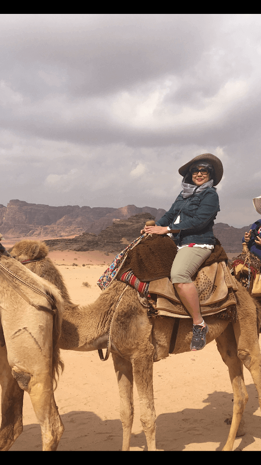 Desert adventure with woman riding camel in scenic landscape, wearing hat and sunglasses, under cloudy sky.