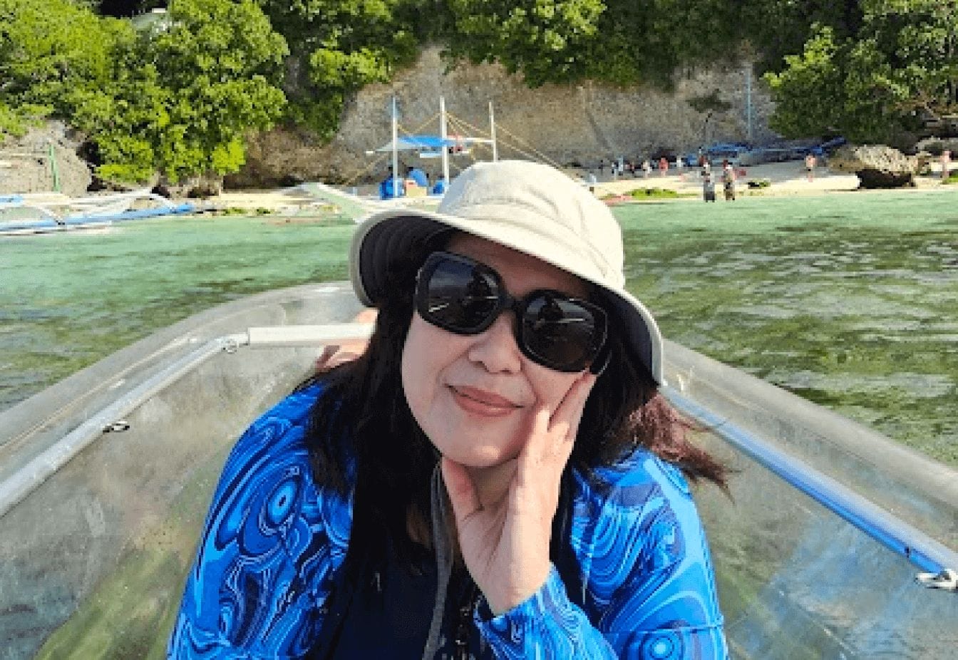 Woman in sunglasses and hat on a boat, enjoying a sunny day near a tropical beach with clear water and lush greenery.